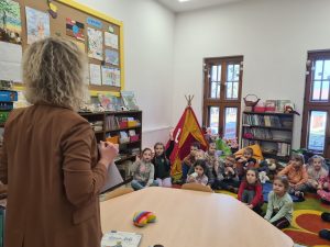 Dzieci siedzą na dywanie w dziale dziecięcym i udzielają odpowiedzi na zagadki zadane przez bibliotekarkę.