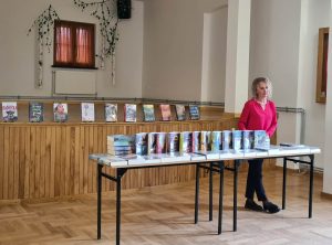 Książki z dużą czcionką są ułożone na stolikach za stolikiem stoi bibliotekarka