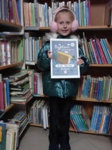 Dziewczynka stoi przed regałem z książkami w rękach trzyma Dyplom Małego Czytelnika