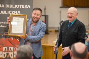 Dwóch rozbawionych mężczyzn stoi przed grupą ludzi. Jeden z nich trzyma w rękach ramę z uchwałą miasta. 