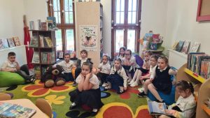 Grupa dzieci siedzi na podłodze w bibliotece. Słuchają czytanej przez bibliotekarkę bajki.