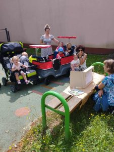 Dzieci siedzą w wózku przed ławką, na której pani czyta bajkę w teatrzyku kamishibai. 