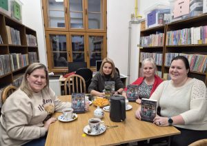 Cztery kobiety siedzą przy stoliku, w dłoniach trzymają książki Barbary Wysoczańskiej "Obiecaj, że wrócisz". Na stole leżą filiżanki z herbatą. 