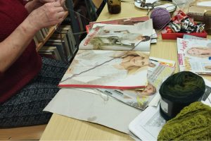 Na stole leżą czasopisma ze ściegami. Obok stołu widać ręce kobiety, która robi coś na szydełku.