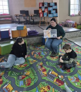 Bibliotekarka pokazuje dzieciom strony w książce, które ilustrują czytany tekst.